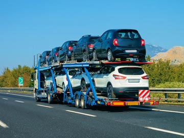 Ein Autotransporter fährt auf einer Autobahn und transportiert mehrere Fahrzeuge, während ein klarer blauer Himmel und grüne Hügel im Hintergrund zu sehen sind.